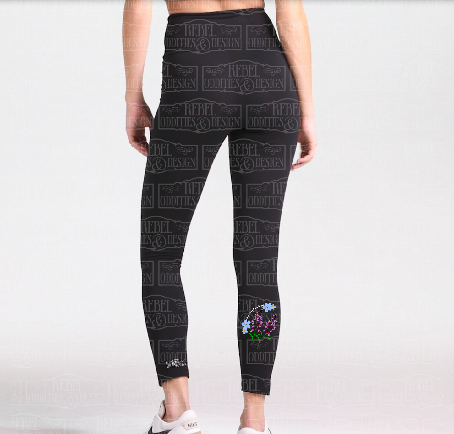 Back view of black leggings, the right leg has a forget-me-not and fireweed design on the calf and a small version of the logo above the ankle seam on the left leg.