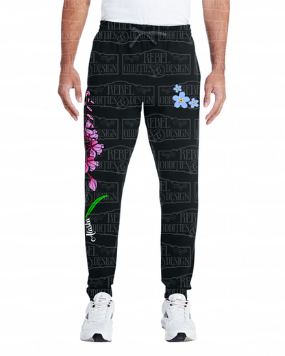Front view of Black jogger sweatpants with a fireweed design up the side of the right leg, featuring script reading 'Alaska' and three forget-me-nots below the left pocket, along with a small version of the logo on the back of the left leg, all on a white background.